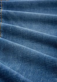 Tejido de mezclilla en un tono azul medio con una textura distintiva, un patrón sutil y costuras visibles a lo largo de los bordes.