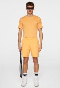 Oranžs sporta krekls un šortu komplekts, izgatavots no viegla auduma. Iekļauts logotips uz krūšu. Valkāts ar baltām kedām un tenisa raketi.