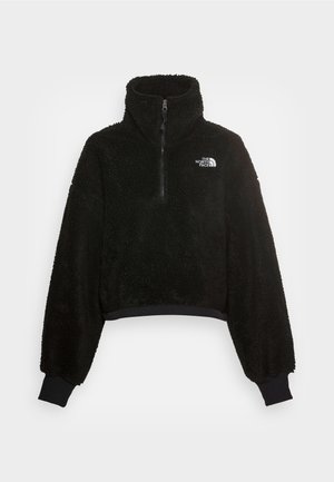 Sudadera de forro polar negra con un alto cuello, cremallera frontal y puños acanalados, que presenta un logo en el lado izquierdo del pecho. Superficie suave y texturizada.