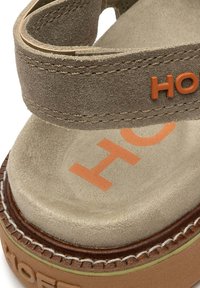 Beige suède platform sandaal met een bruine zool, voorzien van een oranje logo en stikseldetails. Het ontwerp heeft een brede band over de bovenkant.