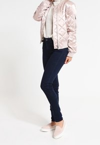 Lichtroze gewatteerd bomberjack, witte trui, donkere strakke jeans en roze instapschoenen met witte zolen. Gladde texturen en modern design.