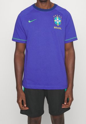 T-shirt Nike viola con logo del Brasile e stelle, con dettagli verdi sulle maniche. Abbinata a pantaloni corti neri, che mostrano un design texture.