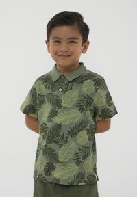 Polo a maniche corte di colore verde oliva con stampa di foglie in verde chiaro e verde scuro. Presenta un colletto e due bottoni frontali.