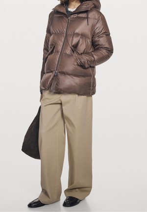 Persoon draagt een bruine donsjas, beige broek met wijde pijpen, zwarte schoenen en draagt een donkerbruine tas, staand tegen een effen achtergrond.