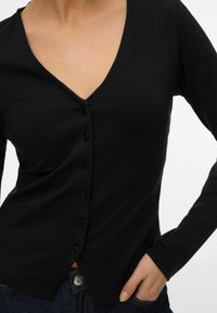 Femme portant un haut noir ajusté à manches longues avec boutons et col en V, main dans la poche d'un jean bleu foncé, torse visible sur fond blanc.