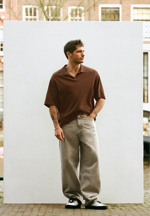 Mężczyzna z tatuażami, ubrany w brązową koszulkę polo, luźne szare dżinsy i trampki, stojący na tle jednolitego białego tła na zewnątrz.