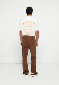 Camiseta blanca con estampado gráfico naranja y pantalones de pana marrones. Zapatillas marrones con detalles blancos. Diseño simple, ajuste estándar.