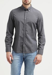 Tummanharmaa pitkähihainen napitettava paita, jossa on hienovarainen kuvio, kaulus ja yksi rintatasku, yhdistetty sinisiin farkkuihin.