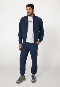 Tamsiai mėlynas vėjaraštis su nertomis rankogaliais ir apačia, derinamas su atitinkamomis kelnėmis, turinčiomis subtilų tekstūrinį raštą ir kišenes. Pilki sportiniai batai.