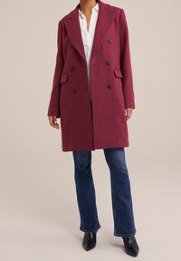 Cappotto doppiopetto in misto lana color bordeaux con rever a lancia, tasche frontali e bottoni neri, abbinato a jeans svasati e stivaletti alla caviglia.