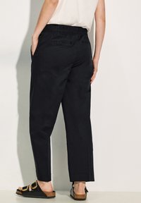 Pantaloni neri con cintura elastica e vestibilità ampia, dotati di tasche laterali e orli a cuffia. Indossati con sandali neri e una maglietta di colore chiaro.