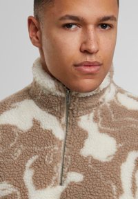 Pullover in pile marrone e bianco con zip a metà, dotato di un colletto alto, caratterizzato da un motivo testurizzato e dettaglio in argento sulla zip del collo.