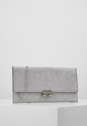 Pochette argentée texturée avec une bandoulière en chaîne argentée et un fermoir argenté rectangulaire posée sur une surface blanche.