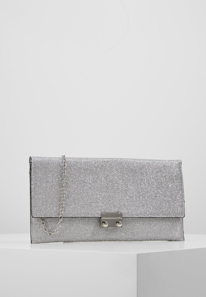 Pochette argentée texturée avec une bandoulière en chaîne argentée et un fermoir argenté rectangulaire posée sur une surface blanche.
