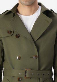 Trinchera de color verde oliva de doble botonadura con grandes botones marrones, cinturón en la cintura y cuello puntiagudo, hecha de un tejido suave.