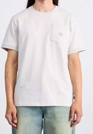 T-shirt gris clair en coton à manches courtes avec une poche poitrine et un logo brodé. Texture lisse et coupe décontractée.