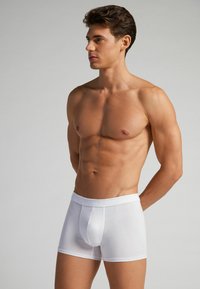 Weiße Baumwoll-Boxershorts mit einem enganliegenden Design und elastischem Bund, ausgestattet mit einer Fronttasche und nahtlosen Seiten. Glatte Textur.