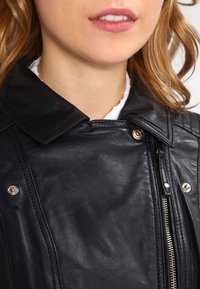 Černá kožená bunda s pevně tvarovaným límcem, stříbrnými kovovými prvky a viditelnými detaily zipu. Hladká textura s jemnými švy.