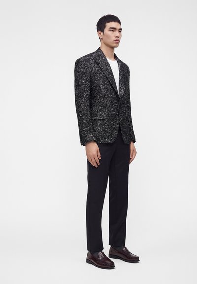 Homme debout portant un blazer texturé noir et blanc, une chemise blanche, un pantalon noir et des mocassins marron foncé sur fond blanc uni.