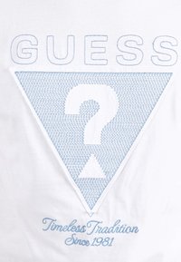 Camiseta blanca con un logo triangular bordado en azul que tiene un signo de interrogación. El texto dice "GUESS" arriba y "Tradición Intemporal Desde 1981" abajo.