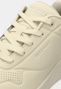 Ανοιχτό μπεζ αθλητικό παπούτσι με λείο, υφασμένο δερμάτινο μέρος επάνω, επίπεδα κορδόνια και διακριτικό λογότυπο. Διαθέτει διάτρητα στοιχεία στο πλάι.