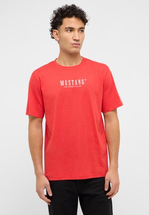 Junger Mann mit lockigem Haar, der ein rotes Mustang-T-Shirt und schwarze Hosen trägt, vor einem schlichten hellen Hintergrund stehend, zur Seite blickend.
