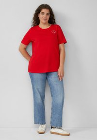 Rotes Baumwoll-T-Shirt mit kurzen Ärmeln und einem Herzsymbol auf der linken Brust, kombiniert mit hellblauen geraden Jeans und weißen Sneakers.