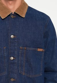 Chaqueta de mezclilla con un cuello de pana marrón, botones plateados, costuras naranjas y un bolsillo en el pecho que presenta una etiqueta de marca.