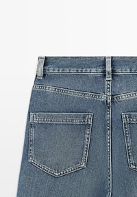 Vista posteriore di jeans in denim blu che mostrano due tasche posteriori, passanti per cintura e cuciture visibili su uno sfondo bianco.