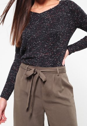 Woman wearing a black speckled long-sleeve sweater tucked into high-waisted olive pants with a tied fabric belt.
