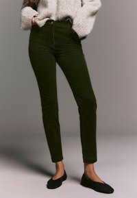 Pantaloni din corduroy verde măsliniu, cu o croială slim, talie înaltă și buzunare frontale, asortați cu un pulover texturat de culoare deschisă și pantofi negri fără toc.