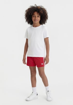 Barn med krøllet hår står iført en enkel hvid t-shirt, røde Adidas-shorts, hvide sokker og hvide sneakers mod en hvid baggrund.