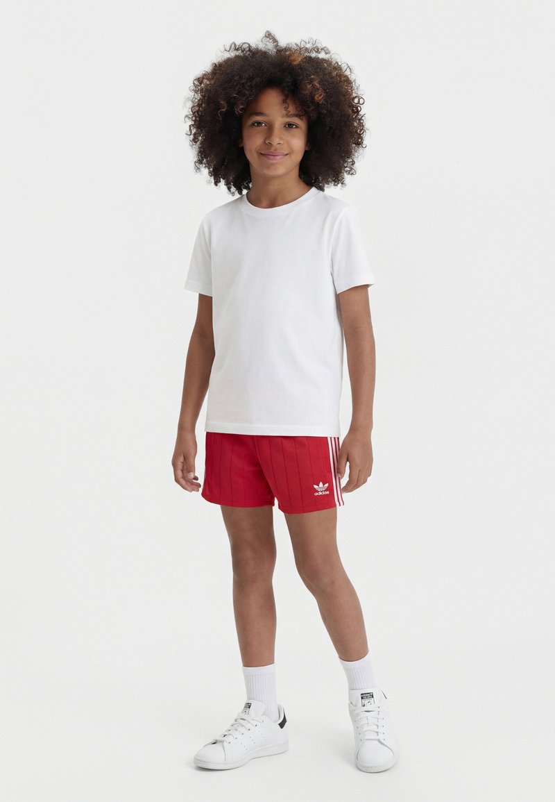 Enfant aux cheveux bouclés debout, portant un t-shirt blanc uni, un short rouge Adidas, des chaussettes blanches et des baskets blanches sur fond blanc.