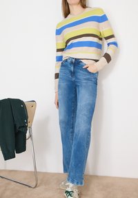 Personne portant un jean large bleu, un pull rayé multicolore et des baskets vertes et blanches, debout à côté d'une chaise avec une veste sombre.