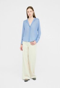 Lyseblå langærmet bluse med krave og knapdetaljer, kombineret med løstsiddende cremefarvede bukser og sorte flade sko.