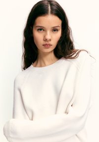 Fehér kötött pulóver bordázott, kerek nyakkivágással és leejtett vállas kialakítással, puha textúrával és laza szabással.