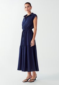 Tamsiai mėlynas maksi suknelė su trumpomis rankovėmis, mygtukų užsegimu priekyje, pabrėžta liemeniu ir kelis sluoksnius turinčiu apvadu, pagaminta iš lengvos medžiagos.