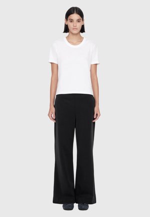 Woman with straight dark hair wearing a plain white short-sleeve shirt, wide black pants, and dark shoes standing against a light background.