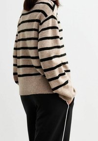 Pull en tricot beige avec des rayures horizontales noires, présentant une coupe décontractée et des poignets côtelés. Associé à un pantalon noir, affichant un look décontracté.