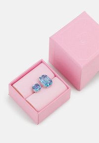 Zwei blaue Edelsteine in silbernen Fassungen befinden sich in einer hellrosa, strukturierten Box mit weicher Polsterung. Der größere Stein ist rechteckig, der kleinere ist quadratisch.