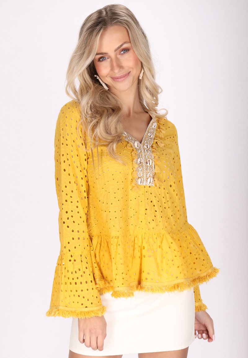 Blusa amarilla con patrón de ojal, mangas acampanadas y dobladillo peplum; presenta un borde decorativo a lo largo del escote y las mangas.