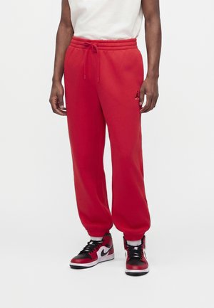 Homme portant un pantalon de jogging rouge avec un cordon et des baskets montantes noires, rouges et blanches, debout devant un fond blanc.