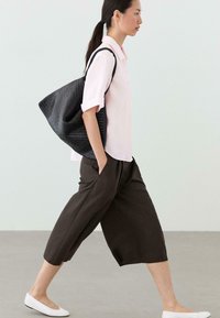 Vrouw loopt met één hand in haar zak, draagt een lichtroze blouse, zwarte wijde cropped broek, witte platte schoenen en draagt een grote zwarte getextureerde schoudertas.