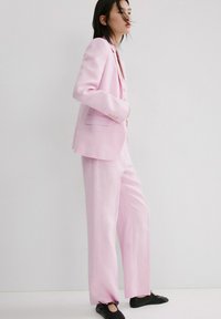 Femme debout portant un tailleur rose clair avec un pantalon à jambes larges et un blazer, associé à des chaussures plates noires, sur un fond uni.