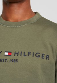 Sudadera verde oliva con cuello redondo y logotipo bordado de "TOMMY HILFIGER". Presenta un pequeño emblema en rojo, blanco y azul. Textura suave.
