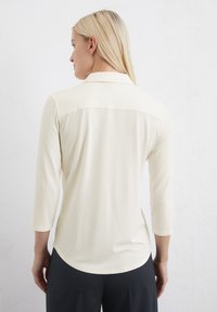 Light cream, long-sleeve polo shirt with a collared neckline, smooth texture, and a curved hem. Features a back yoke for added detail.