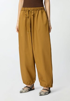 Pantaloni larghi giallo senape con vita e caviglie elastiche, abbinati a ballerine appuntite in pelle di serpente e top marrone senza maniche.