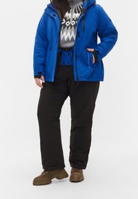 Chaqueta aislante azul con capucha, suéter de patrón gris debajo, pantalones de nieve negros y botas de senderismo marrones con suelas gruesas.