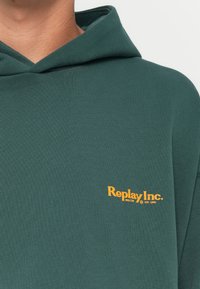 Felpa verde realizzata in un tessuto morbido, dotata di tasca a marsupio e di un piccolo logo giallo "Replay Inc." sul davanti.
