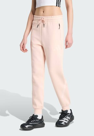 Person iført lyserøde joggingbukser med lynlåslommer, hvid cropped top og sorte og hvide sportssko på lys baggrund.